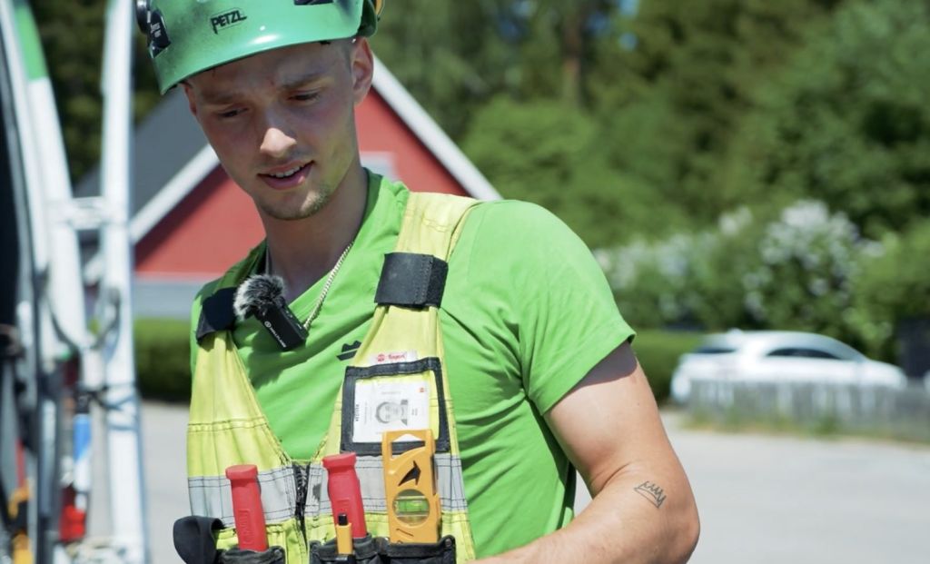 En vesivekanställd klädd i grön hjälm och t-shirt med verktyg i verkstygsbältet. Står framför röd villa där de genomför en takrenovering.