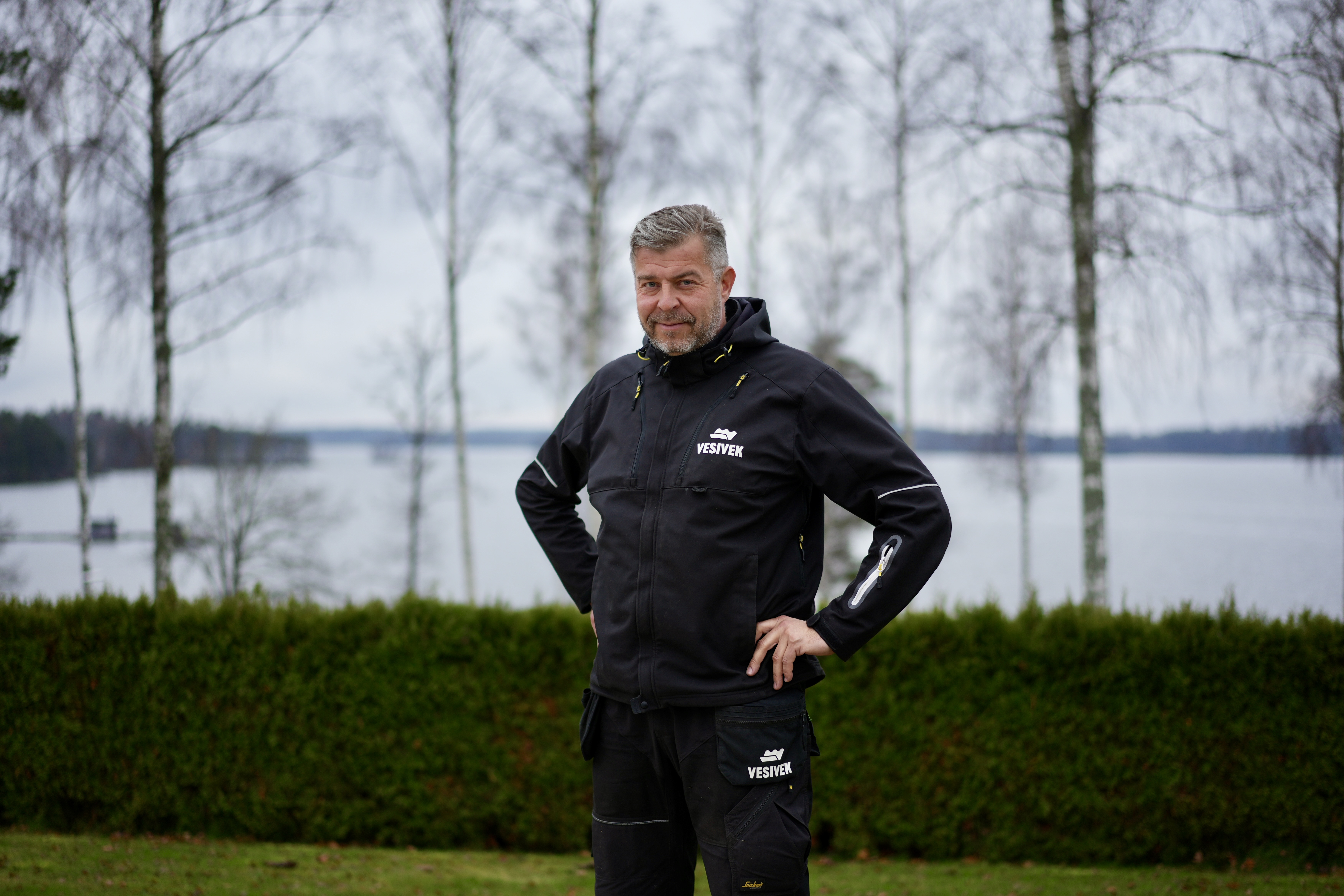 Jani Jylhä Vd på Vesivek står utomhus med vatten och björkar i bakgrunden.